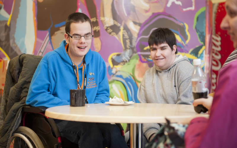 Two South West College students sitting at a table, one is using a wheelchair